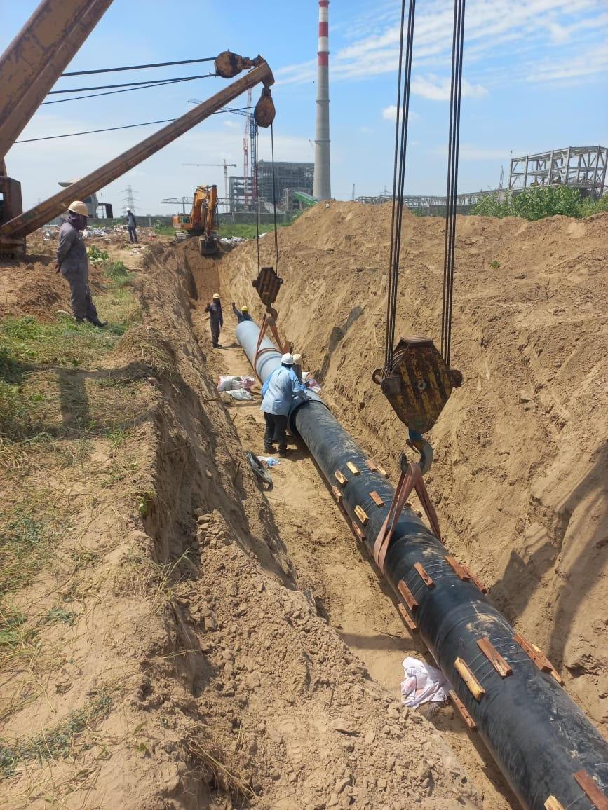 Pipe string lowered into trench with power plant cooling towers in the background